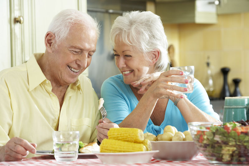 an elderly couple smiling at each other over a meal because denture implants were used to benefit the look and function of their smiles.